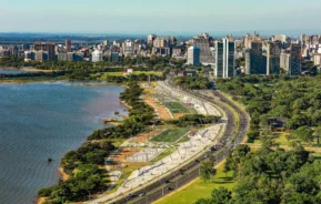 Imagem da vista aérea da orla do Guaíba, em Porto Alegre, mostra a paisagem urbana à beira-mar com arranha-céus modernos, uma linha costeira curva, parques verdes, ruas arborizadas e um grande rio sob um céu claro.