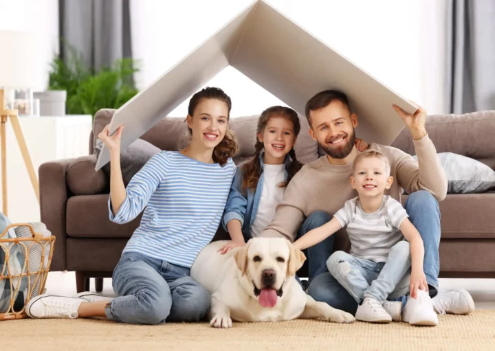 Imagem de uma família composta por um pai, uma mãe, dois filhos crianças e um cachorro sentados no chão da sala com um papelão em cima da cabeça sinalizando o telhado de uma casa para ilustrar matéria sobre como aumentar a segurança em apartamento