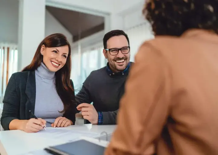 Imagem de um homem e uma mulher sorridentes sentados em frente a outra pessoa em uma mesa, parecendo felizes enquanto discutem em um ambiente de escritório moderno e bem iluminado