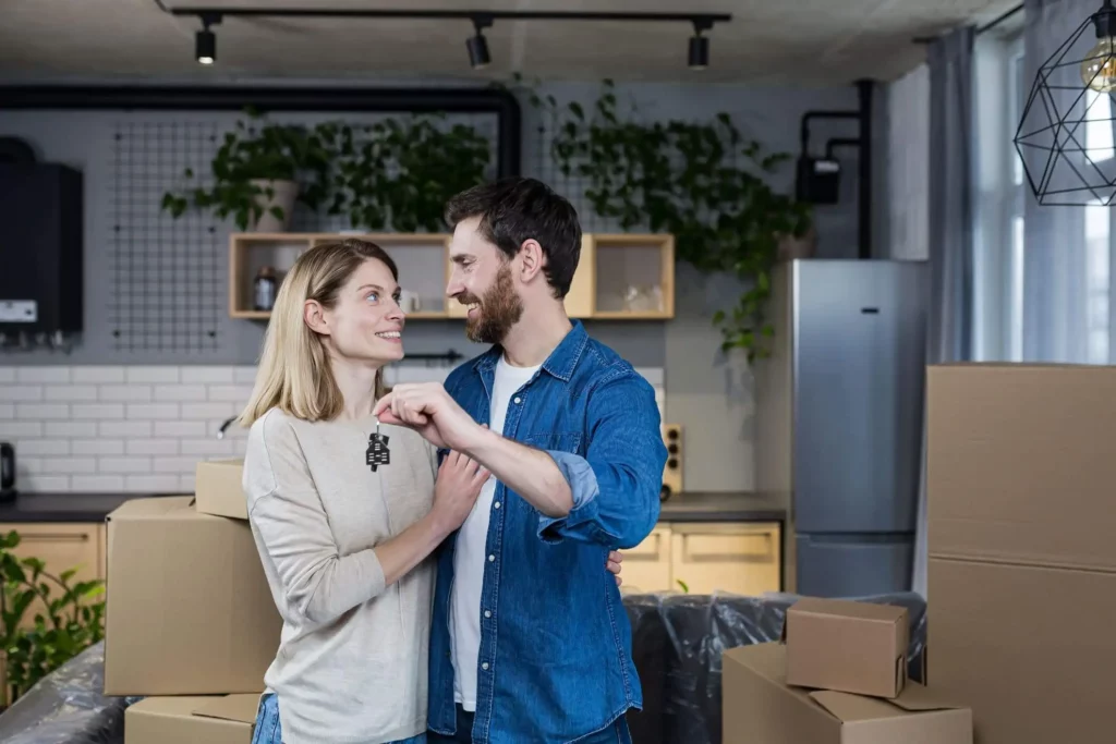 Imagem de um casal abraçados segurando a chave de uma casa em um ambiente cheio de caixas de mudança para ilustrar matéria sobre como financiar imóvel pela Caixa sem carteira de trabalho assinada