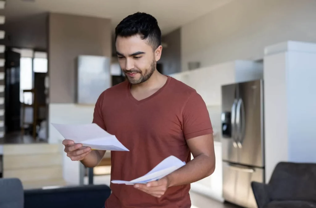Imagem de um homem de cabelos escuros curtos e barba aparada, vestindo uma camiseta cor de ferrugem, lendo jornais em uma cozinha e sala de estar modernas e bem iluminadas para ilustrar matéria sobre como liberar FGTS para compra de imóvel à vista