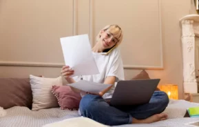 Imagem de uma pessoa sentada de pernas cruzadas em uma cama com um laptop, segurando papéis e falando ao telefone, parecendo ser multitarefa enquanto pesquisa sobre consórcio ou financiamento de casa em um quarto aconchegante e bem iluminado.