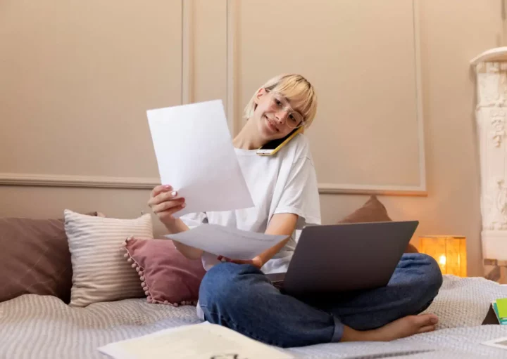 Imagem de uma pessoa sentada de pernas cruzadas em uma cama com um laptop, segurando papéis e falando ao telefone, parecendo ser multitarefa enquanto pesquisa sobre consórcio ou financiamento de casa em um quarto aconchegante e bem iluminado.