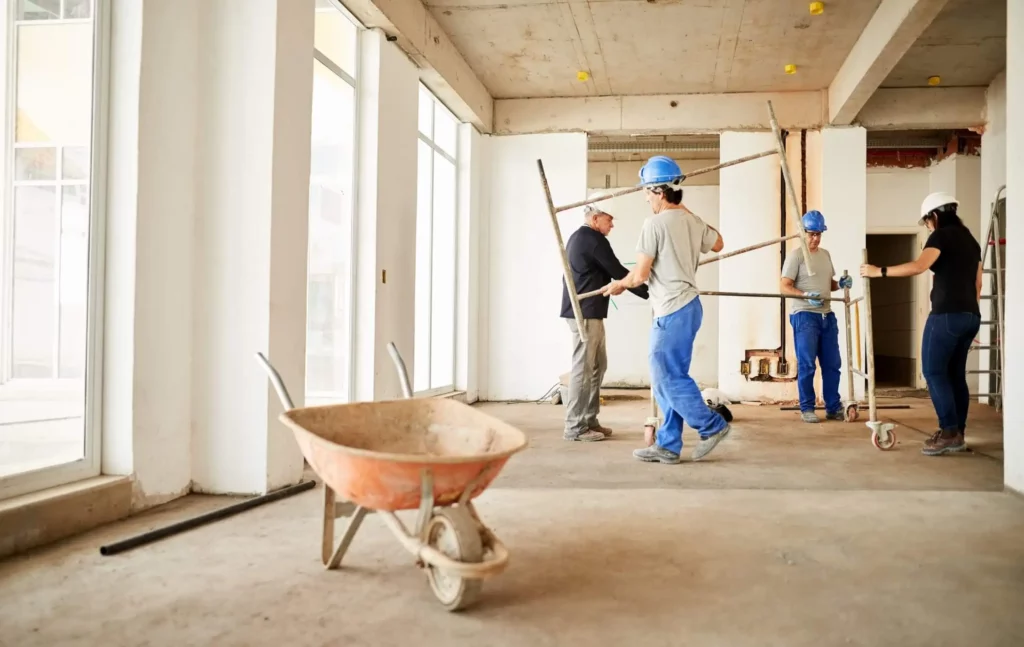 Imagem de trabalhadores em uma casa em obras avaliando a estrutura e carregando ferramentas perto de um carrinho de mão para ilustrar matéria sobre construir ou reformar