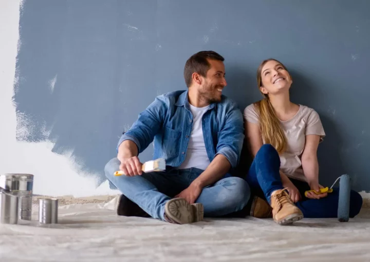 Imagem de um casal composto por um homem e uma mulher sentados no chão de um imóvel ao lado de latas de tinta enquanto ao fundo a parede está quase toda pintada na cor azul para ilustrar matéria sobre construir ou reformar