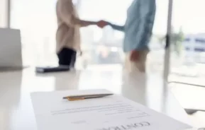 Imagem de um contrato de aluguel sobre uma mesa em foco nítido, enquanto duas pessoas em trajes de negócios apertam as mãos no fundo desfocado, sugerindo um contrato de aluguel ou negócio bem-sucedido