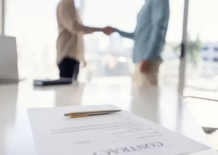 Imagem de um contrato de aluguel sobre uma mesa em foco nítido, enquanto duas pessoas em trajes de negócios apertam as mãos no fundo desfocado, sugerindo um contrato de aluguel ou negócio bem-sucedido