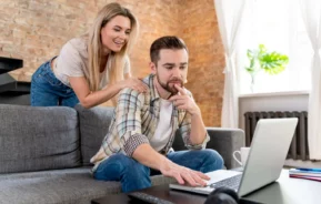 Imagem de um homem sentado em um sofá cinza usando um laptop, parecendo pensativo, para ilustrar matéria sobre crédito imobiliário. Uma mulher está atrás dele, sorrindo e apoiando as mãos em seus ombros. Uma mesa de centro com um controle remoto e fones de ouvido está na frente deles.