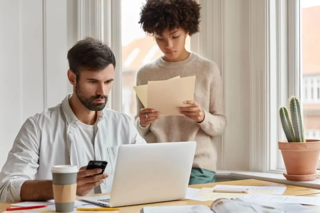 Imagem de um homem sentado em uma mesa usando um laptop e um smartphone para pesquisar como funciona o crédito imobiliário rural, enquanto uma mulher está ao seu lado segurando papéis. A escrivaninha tem uma xícara de café, documentos e um cacto em vaso ao lado de grandes janelas.