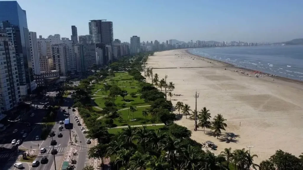 Imagem da vista aérea do Jardim da Orla, em Santos, mostra uma praia urbana com uma ampla faixa de areia, áreas verdes com palmeiras e prédios altos ao longo da orla