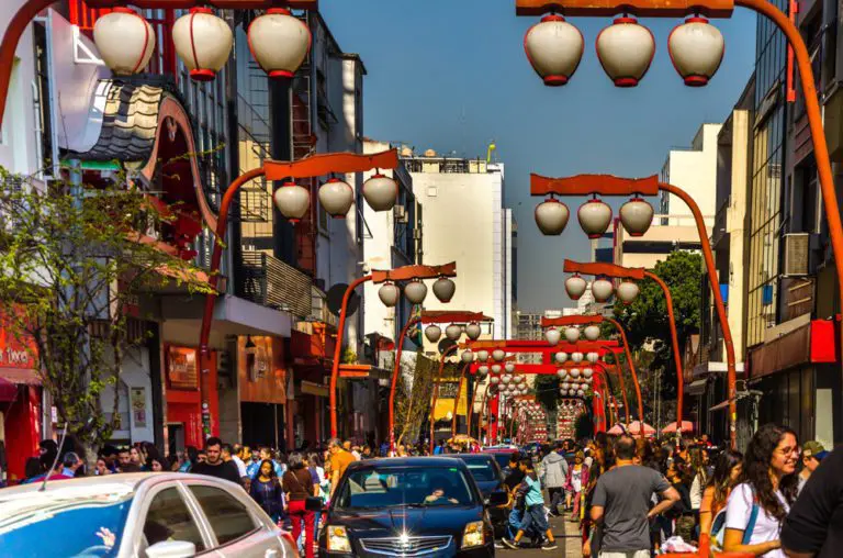 Imagem de rua da Liberdade mostra carros, comércios, visitantes e famosas luminárias em estilo japonês para ilustrar matéria sobre o nome do bairro japonês em São Paulo