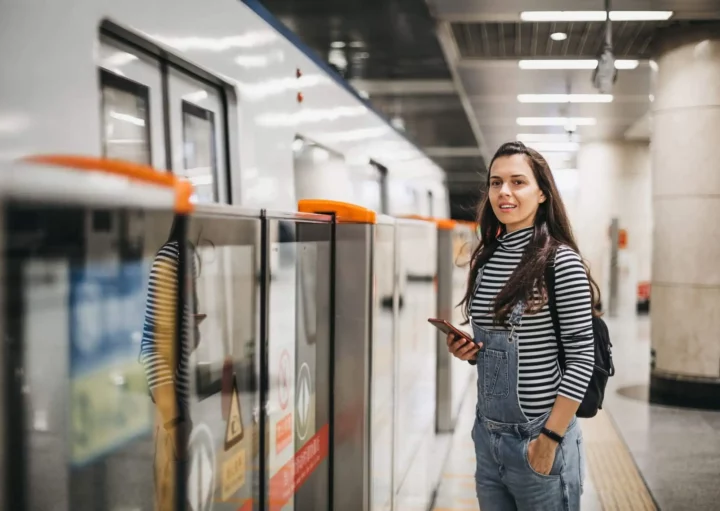 Imagem de uma mulher jovem sorrindo com o celular na mão enquanto aguarda o trem em uma estação de metrô para ilustrar matéria sobre Linha 9-Esmeralda