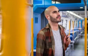Imagem de um homem escutando música com fones de ouvido em um trem, vestindo uma camisa xadrez marrom e carregando uma mochila, para ilustrar matéria sobre a Linha 9-Esmeralda de São Paulo.