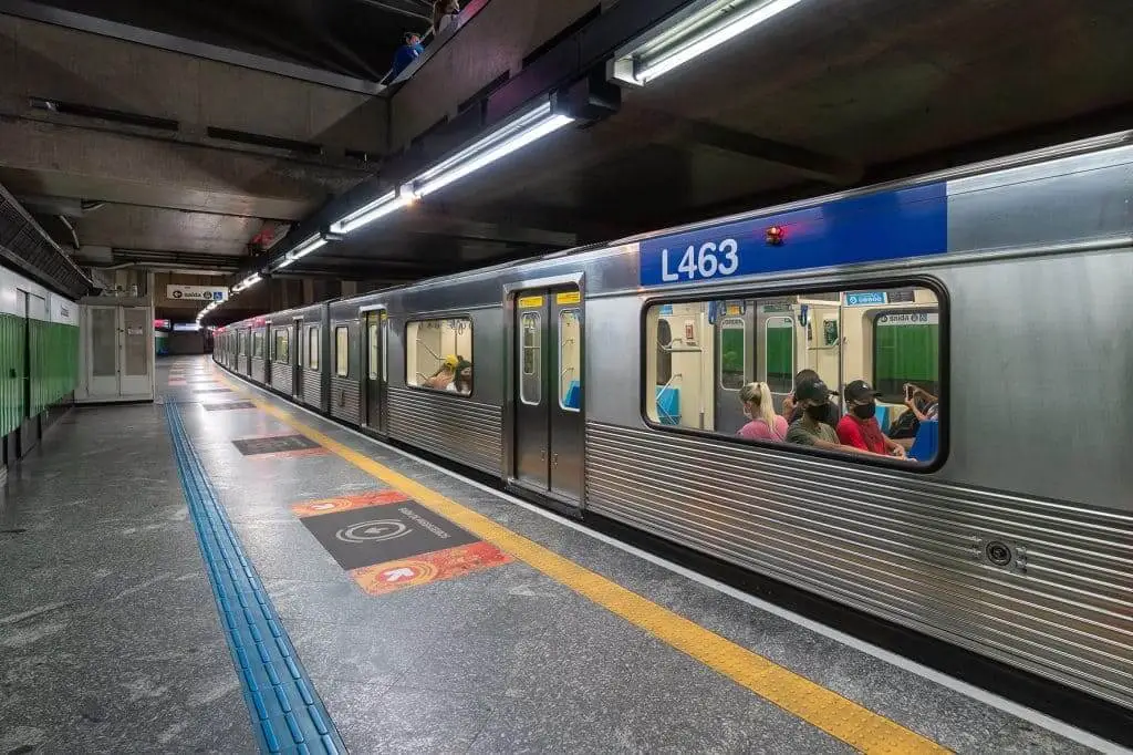 Imagem de um trem de metrô prateado parado na estação Conceição, da Linha Azul do metrô SP. Vários passageiros sentados podem ser vistos através das janelas. 