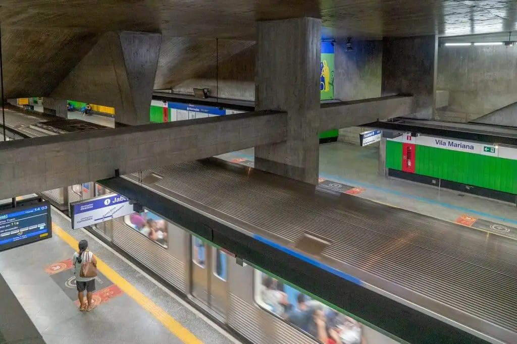 Uma imagem da estação Vila Mariana, da Linha Azul do metrô SP, com vigas de concreto, um trem chegando à plataforma e alguns passageiros esperando. Azulejos verdes e azuis revestem as paredes.