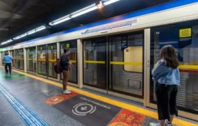 Imagem de pessoas na estação Jabaquara, da Linha Azul do metrô SP, de frente para as portas fechadas do trem. As marcações e os sinais de segurança são visíveis no piso e nas portas da plataforma.