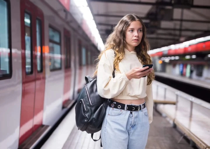 Imagem de uma mulher em uma estação de metrô com um celular na mão na frente de um trem para ilustrar matéria sobre a Linha 3-Vermelha do metrô de SP