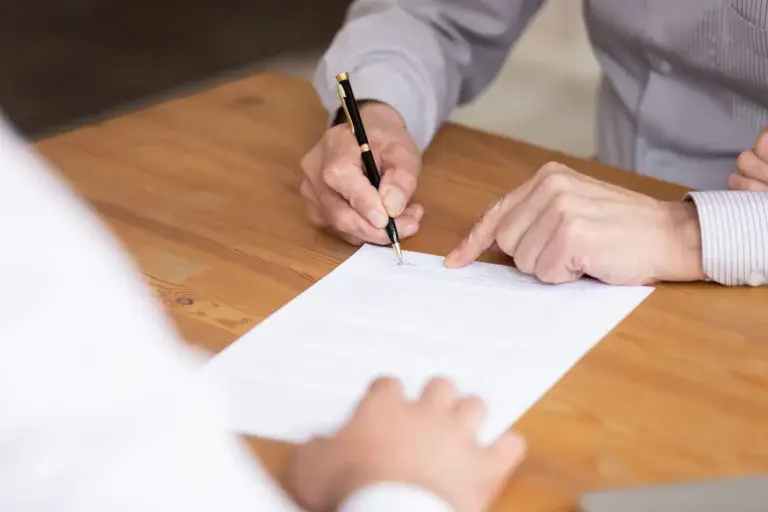 Imagem de duas pessoas sentadas em uma mesa de madeira; uma está assinando um contrato de aluguel residencial enquanto a outra aponta para uma seção do papel. Ambos, vestidos com camisas de mangas compridas, parecem concentrados enquanto finalizam o contrato de aluguel