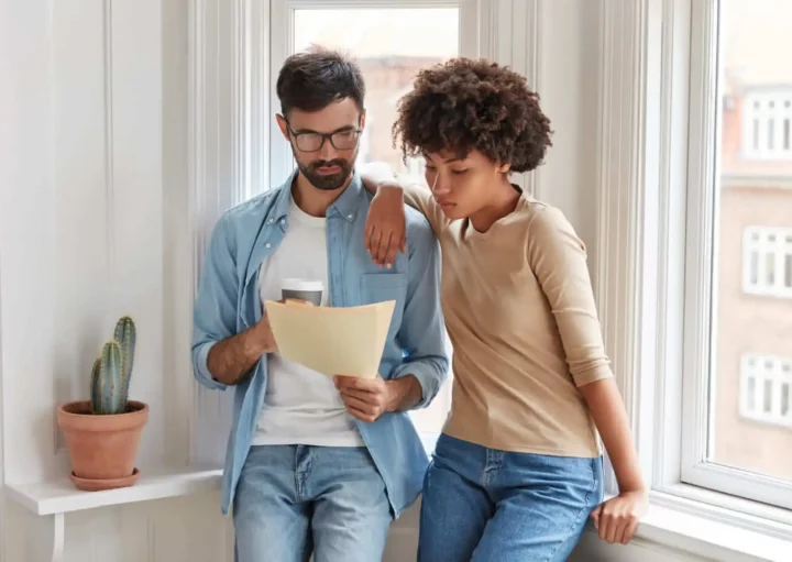 Imagem de um casal formado por um homem e uma mulher em pé encostados na janela de uma casa olhando para documentos que estão na mão do homem para ilustrar matéria sobre o que é usucapião