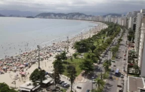 Imagem da vista aérea da orla e uma praia em Santos. A praia está lotada com guarda-sóis, banhistas no mar e um calçadão arborizado, além de uma rua movimentada ao lado de prédios altos sob um céu nublado.