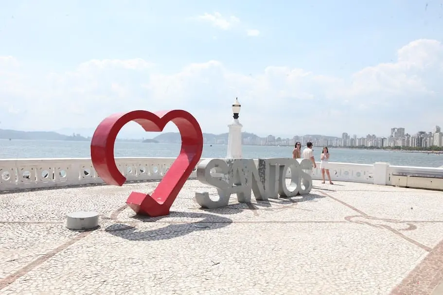 Imagem de um letreiro com um grande coração vermelho e letras cinzas que formam a palavra “SANTOS” se destacam em um calçadão à beira-mar