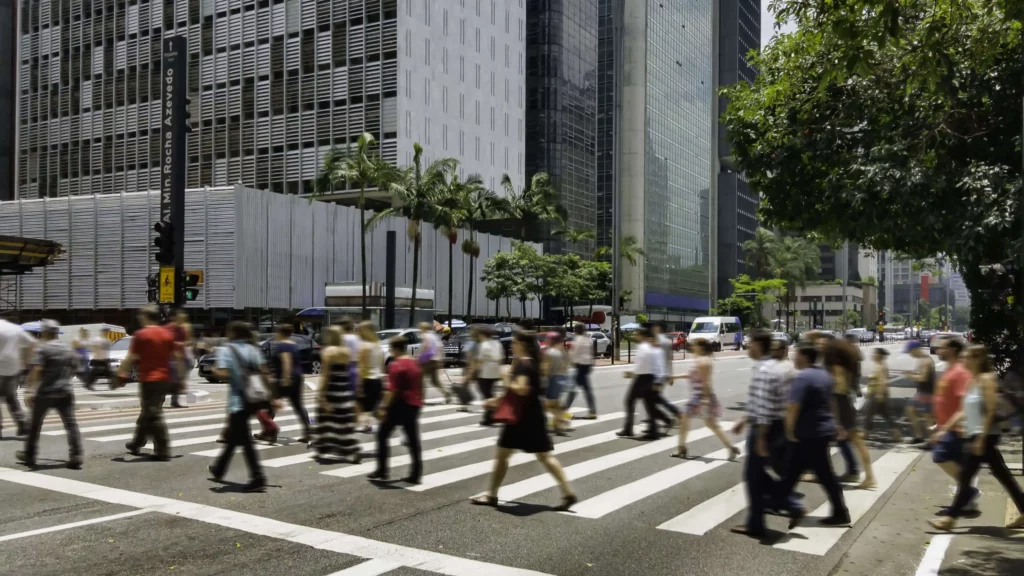 Imagem de pessoas em movimento atravessando uma faixa de pedestre em São Paulo para ilustrar matéria sobre quais os bairros mais populosos de SP