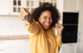 Imagem de uma pessoa sorridente de camisa amarela segura as chaves de casa com um chaveiro em forma de casa em pé em uma cozinha iluminada para ilustrar matéria sobre as regras para uso do FGTS para compra de imóvel. A imagem está focada nas chaves, com a pessoa desfocada no plano de fundo