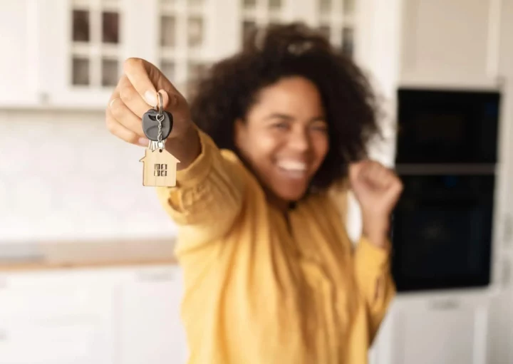 Imagem de uma pessoa sorridente de camisa amarela segura as chaves de casa com um chaveiro em forma de casa em pé em uma cozinha iluminada para ilustrar matéria sobre as regras para uso do FGTS para compra de imóvel. A imagem está focada nas chaves, com a pessoa desfocada no plano de fundo