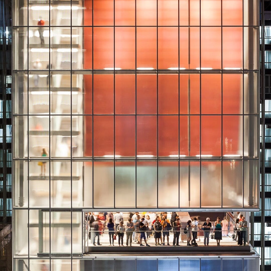 Um grupo de pessoas está em uma varanda bem iluminada dentro de um moderno edifício de vidro perto do metrô Consolação à noite, com uma parede laranja brilhante ao fundo e uma escada visível no lado esquerdo.
