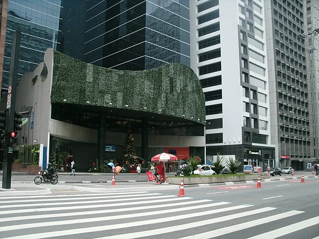 Uma rua moderna perto do metrô Consolação apresenta altos prédios comerciais, um deles com hera verde na fachada. Uma árvore de Natal e enfeites de Natal ficam na entrada, enquanto pessoas, uma motocicleta e um vendedor com um guarda-chuva vermelho se aglomeram na faixa de pedestres.