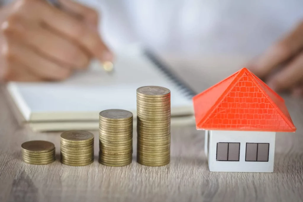 Imagem de pilhas de moedas dispostas em ordem crescente ao lado de uma pequena casa modelo com telhado vermelho. No fundo desfocado, uma pessoa escreve em um caderno