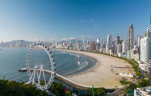 Imagem da vista aérea da praia de Balneário Camboriú mostra faixa de areia, mar, vegetação, roda gigante e construções para ilustrar matéria sobre as cidades que mais crescem em Santa Catarina