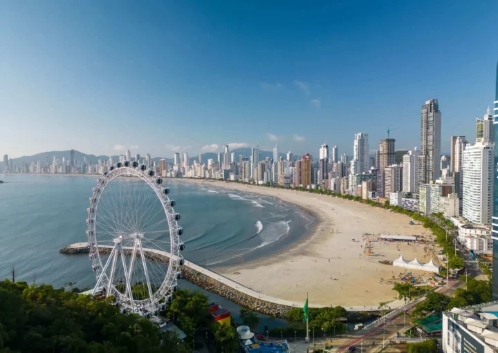 Imagem da vista aérea da praia de Balneário Camboriú mostra faixa de areia, mar, vegetação, roda gigante e construções para ilustrar matéria sobre as cidades que mais crescem em Santa Catarina