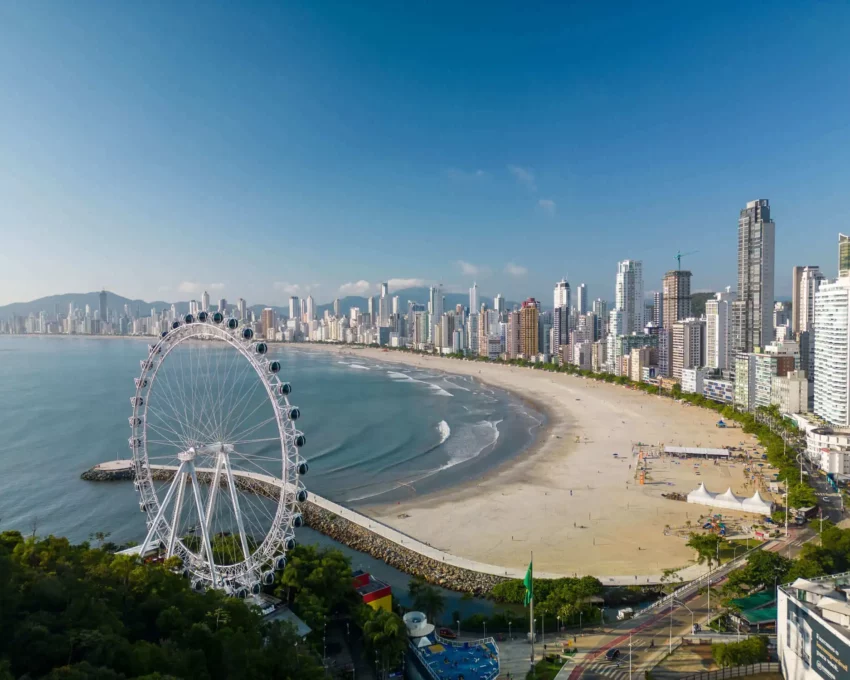 Imagem da vista aérea da praia de Balneário Camboriú mostra faixa de areia, mar, vegetação, roda gigante e construções para ilustrar matéria sobre as cidades que mais crescem em Santa Catarina