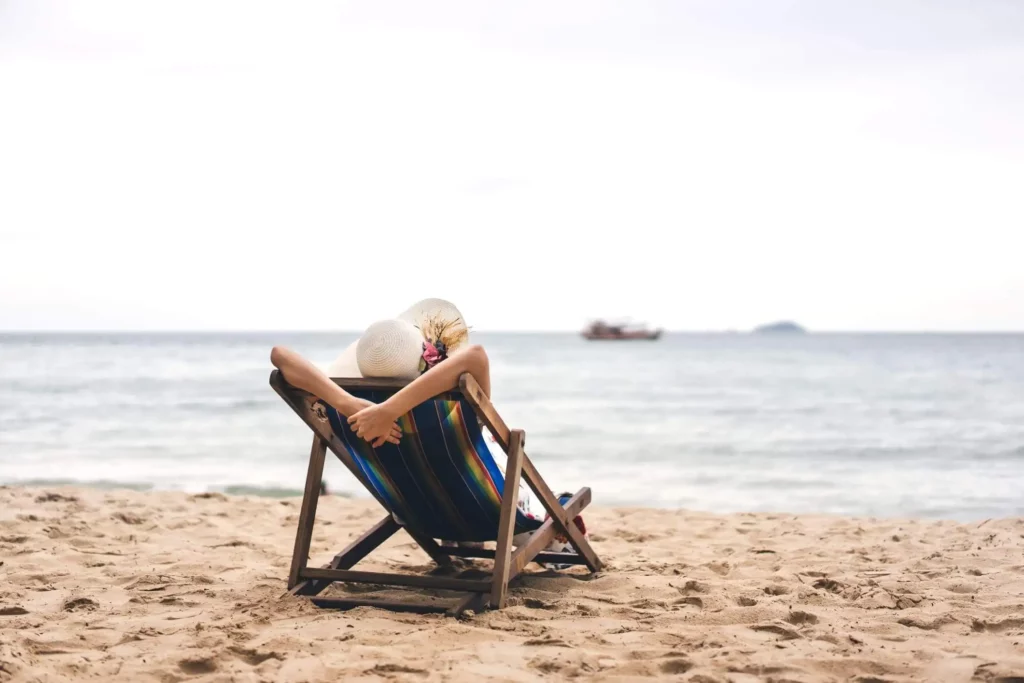 Imagem de uma mulher de costas sentada em uma cadeira espreguiçadeira de praia em cima da faixa de areia olhando o mar para ilustrar matéria sobre as cidades que mais cresceram em Santa Catarina