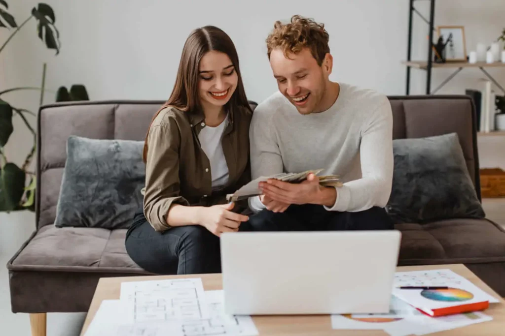 Imagem de um casal sorridente está sentado em um sofá em sua aconchegante sala de estar, analisando juntos os documentos do ITBI. Um laptop e papéis estão espalhados na mesa de centro em frente a eles.
