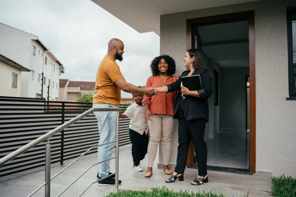 Imagem de um corretor de imóveis apertando a mão de um homem em frente a uma casa, enquanto sua família sorri nas proximidades