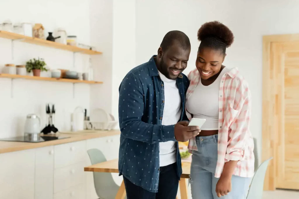 Imagem de um casal sorridente em uma cozinha moderna, olhando para um smartphone enquanto busca informações sobre o que é ITBI. O homem segura o telefone enquanto ambos parecem felizes e engajados, cercados por armários e prateleiras de cores claras.