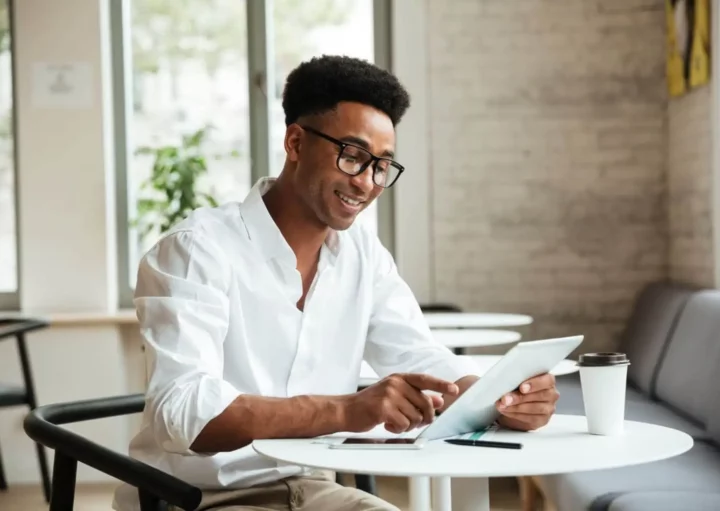 Imagem de um homem de óculos e camisa branca sorrindo enquanto usa um tablet em uma pequena mesa em um café moderno e bem iluminado para ilustrar matéria sobre o que é ITBI. Um smartphone, um notebook e um copo de café descartável estão sobre a mesa.