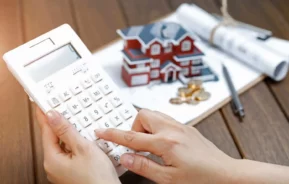 Imagem de uma pessoa usando uma calculadora branca em uma mesa de madeira com uma casa modelo, moedas, caneta e documentos enrolados ao fundo