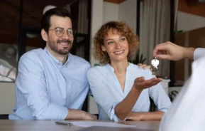 Imagem de um casal sentado à mesa com documentos em cima, recebendo uma chave com chaveiro de casa para ilustrar matéria sobre o que é cessionário de imóvel