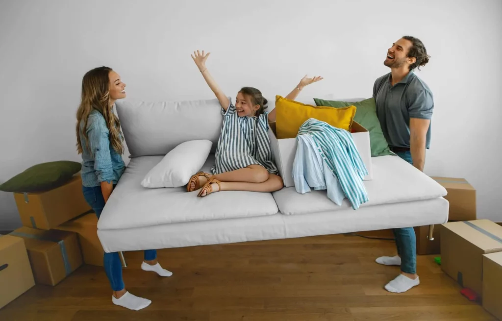 Imagem de um casal sorridente levantando um sofá branco com uma menina risonha sobre ele, cercado de caixas, enquanto se mudam para uma casa nova
