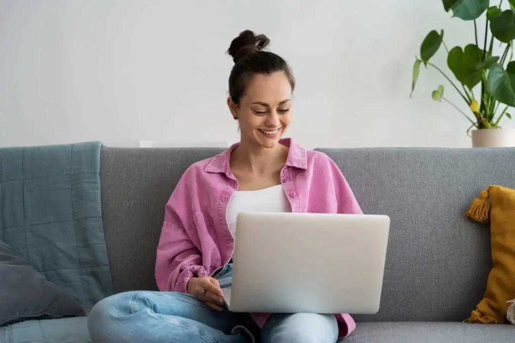 Imagem de uma jovem mulher sentada em um sofá cinza, sorrindo enquanto pesquisa o que é ITBI e trabalha em seu laptop. Vestida com uma camiseta rosa sobre uma blusa branca e jeans azul, ela está cercada por almofadas e uma planta ao fundo.