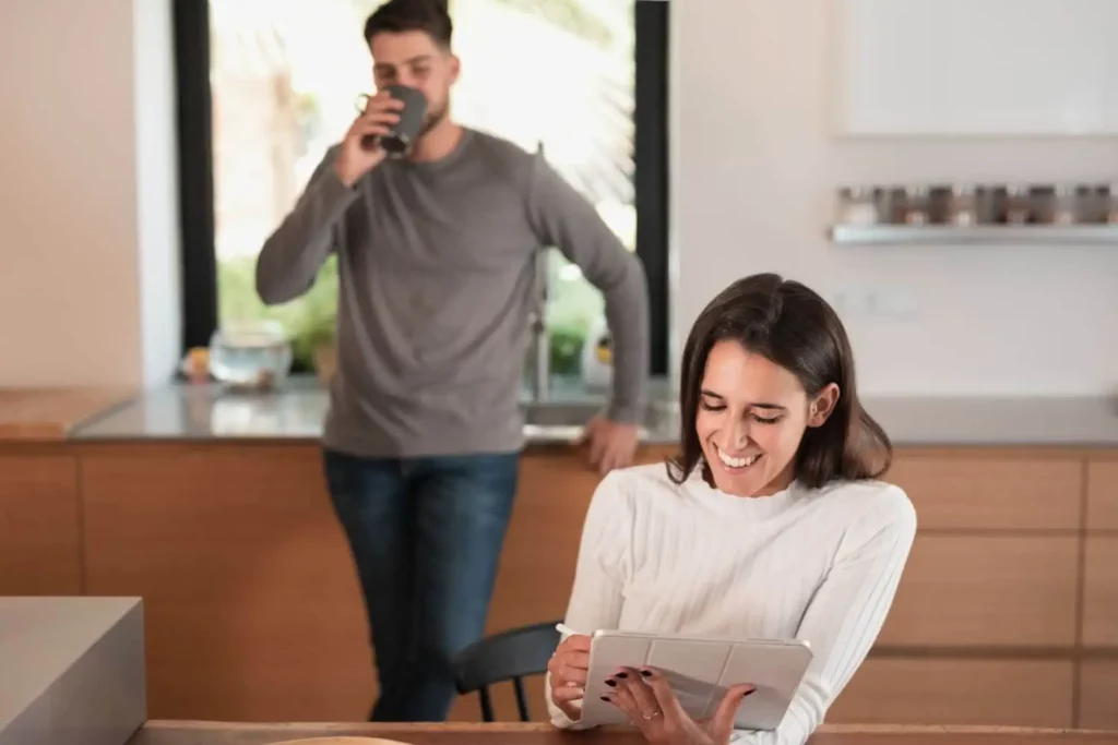 Imagem de uma mulher sentada à mesa da cozinha sorrindo enquanto usa um tablet. Ao fundo, um homem encosta-se ao balcão com uma caneca. A cozinha moderna tem armários de madeira e uma grande janela.