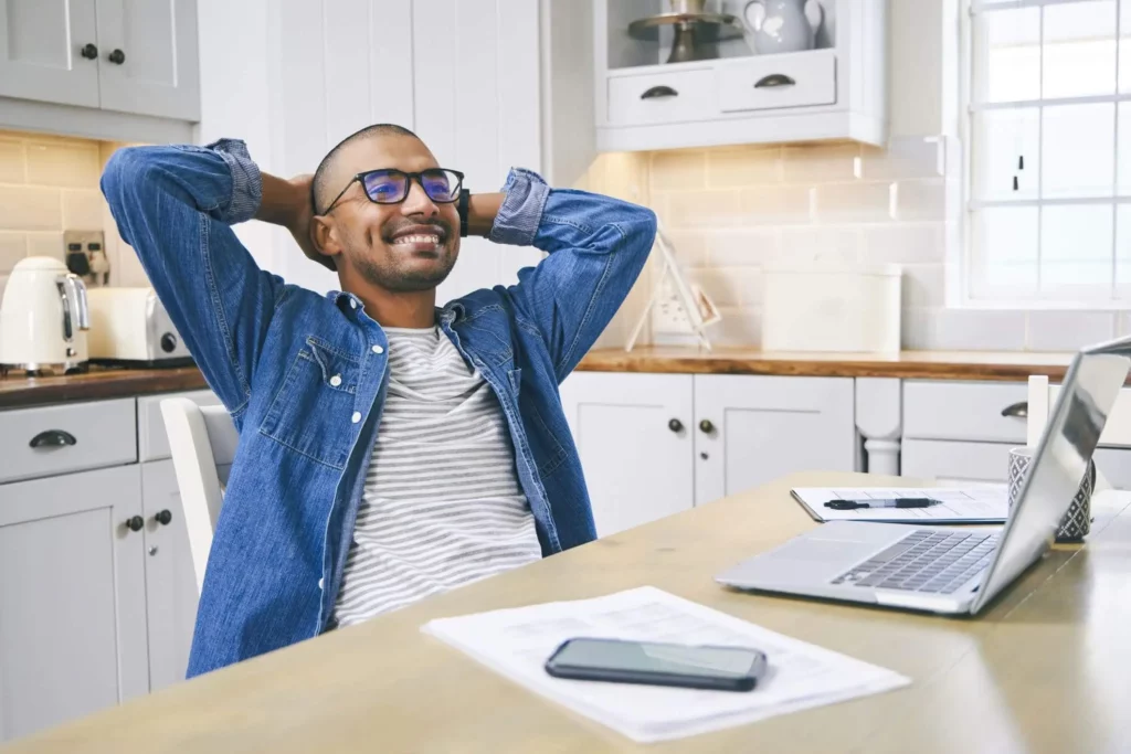 Imagem de um homem sorridente sentado à mesa da cozinha. À sua frente, há um laptop, papéis, um smartphone e uma caneta. A cozinha bem iluminada tem armários brancos e uma janela