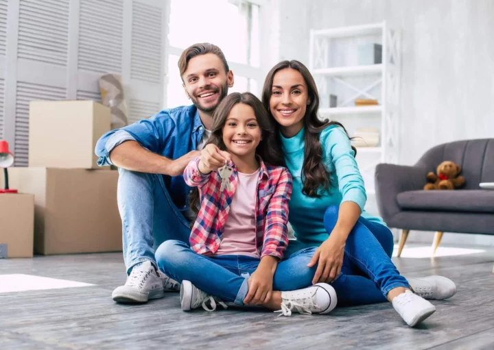 Imagem de uma família sorridente de três pessoas cercada por caixas de mudança comemorando em sua nova sala de estar