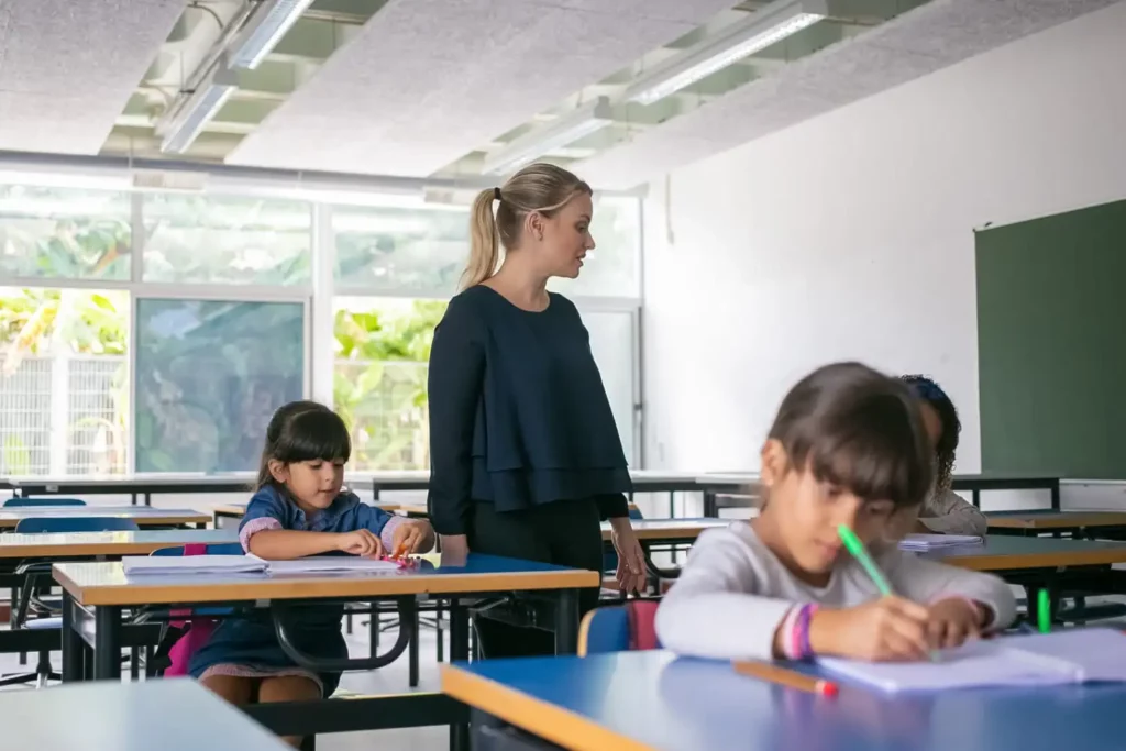 Imagem de uma professora andando entre carteiras escolares enquanto as crianças escrevem em uma folha de papel para ilustrar matéria sobre cidades do Brasil com a melhor educação