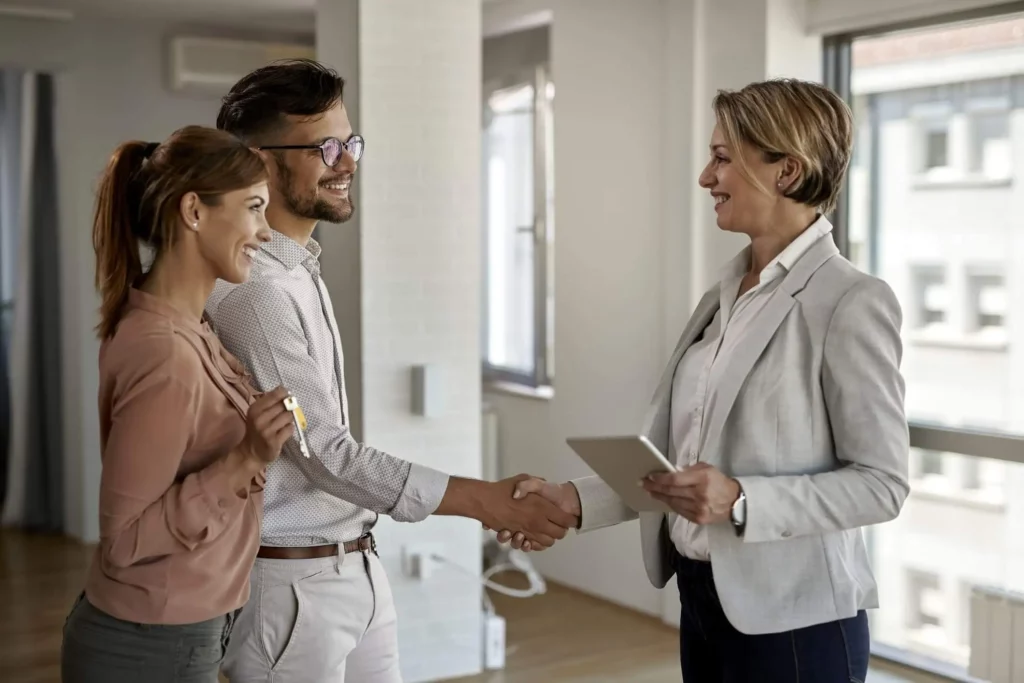 Imagem de um casal de homem e mulher apertando a mão de uma mulher que trabalha no mercado imobiliário dentro de um cômodo de uma propriedade para ilustrar matéria sobre o que é aquisição de imovel