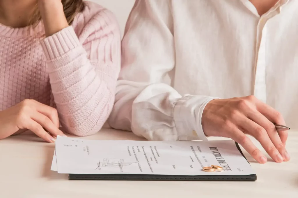 Imagem da silhueta de um homem e uma mulher sentados em frente a uma mesa onde estão papéis de divórcio e duas alianças para ilustrar matéria sobre o que é a partilha de imóvel financiado