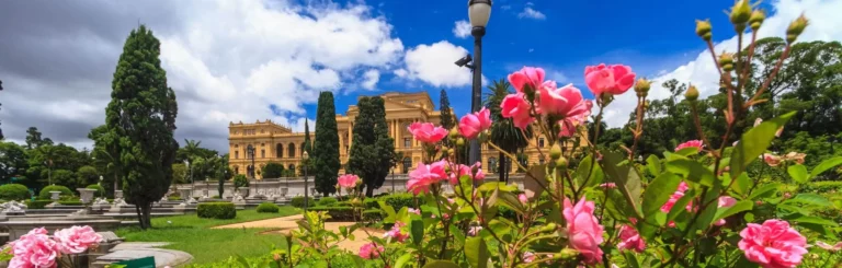 Imagem da vista externa do Parque Independência mostra flores, árvores, vegetação e um edifício em um dia de céu azul para ilustrar matéria sobre parques na Zona Sul de SP